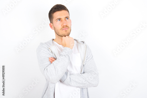 young caucasian man wearing casual clothes over white background  with hand under chin and looking sideways with doubtful and skeptical expression, suspect and doubt.