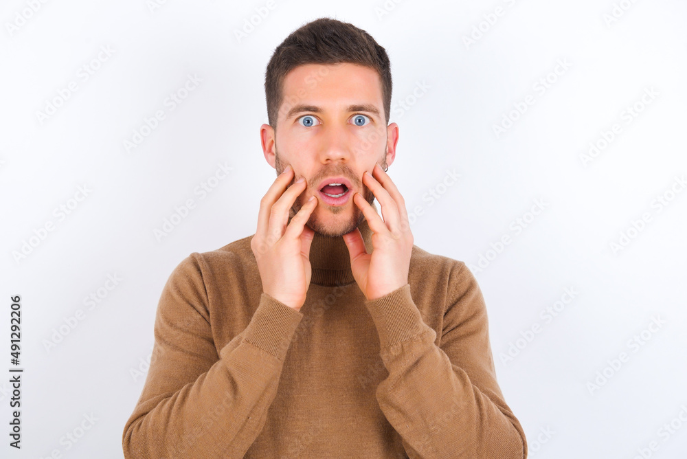 Speechless young caucasian man wearing knitted turtleneck over white background keeps hands near opened mouth reacts to shocking news stares wondered at camera Speechless young caucasian man wearing knitted turtleneck over white background keeps hands near opened mouth reacts to shocking news stares wondered at camera