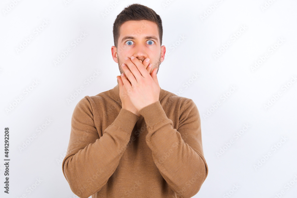 Stunned young caucasian man wearing knitted turtleneck over white background covers mouth with both hands being afraid from something or after hearing stunning gossips. Stunned young caucasian man wearing knitted turtleneck over white background covers mouth with both hands being afraid from something or after hearing stunning gossips.