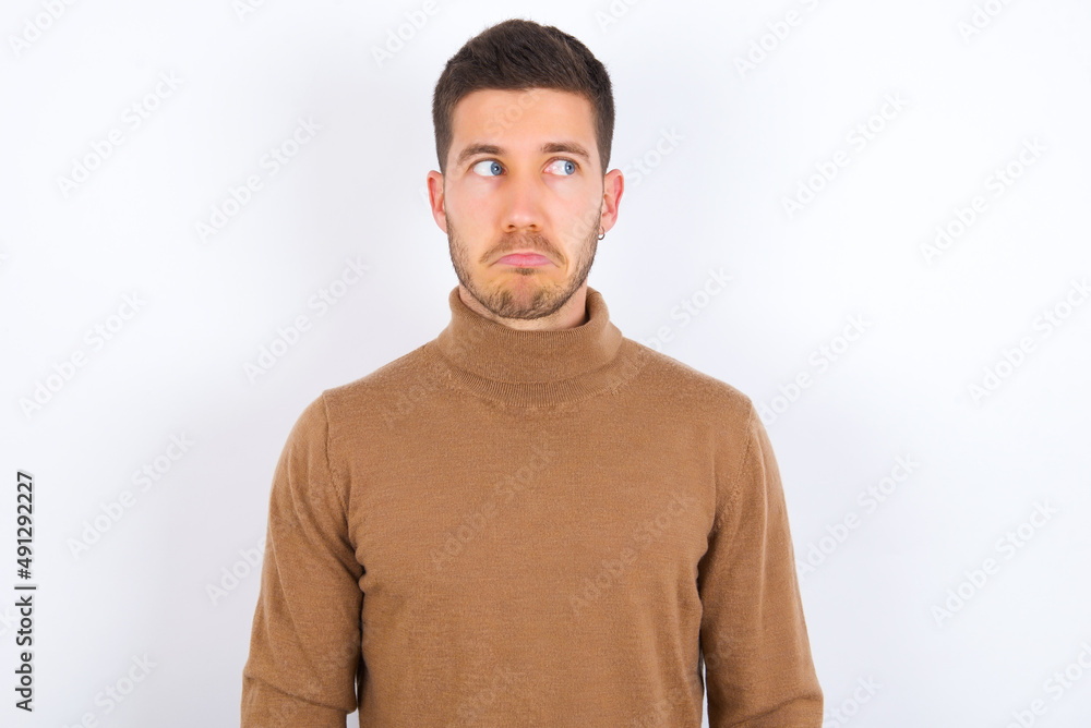 Dissatisfied young caucasian man wearing knitted turtleneck over white background purses lips and has unhappy expression looks away stands offended. Depressed frustrated model. Dissatisfied young caucasian man wearing knitted turtleneck over white background purses lips and has unhappy expression looks away stands offended. Depressed frustrated model.