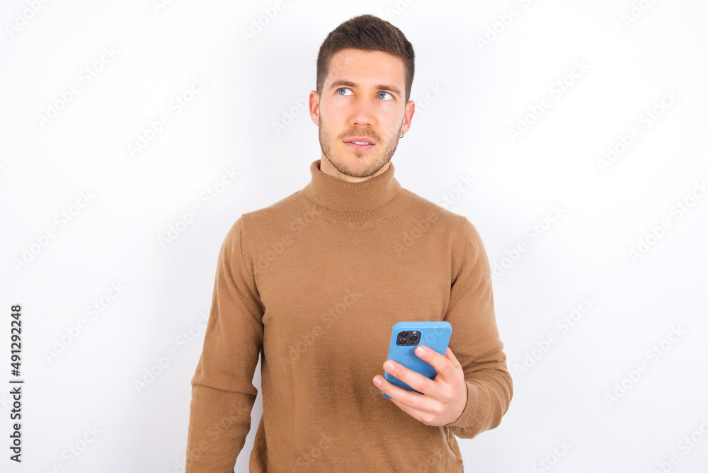young caucasian man wearing knitted turtleneck over white background holds mobile phone uses high speed internet and social networks has online communication. Modern technologies concept young caucasian man wearing knitted turtleneck over white background holds mobile phone uses high speed internet and social networks has online communication. Modern technologies concept