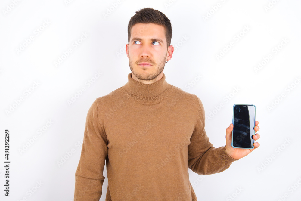 young caucasian man wearing knitted turtleneck over white background holds new mobile phone and looks mysterious aside shows blank display of modern cellular young caucasian man wearing knitted turtleneck over white background holds new mobile phone and looks mysterious aside shows blank display of modern cellular