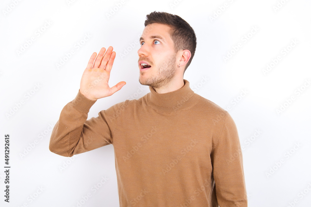 young caucasian man wearing knitted turtleneck over white background look empty space holding hand near her face and screaming or calling someone. young caucasian man wearing knitted turtleneck over white background look empty space holding hand near her face and screaming or calling someone.