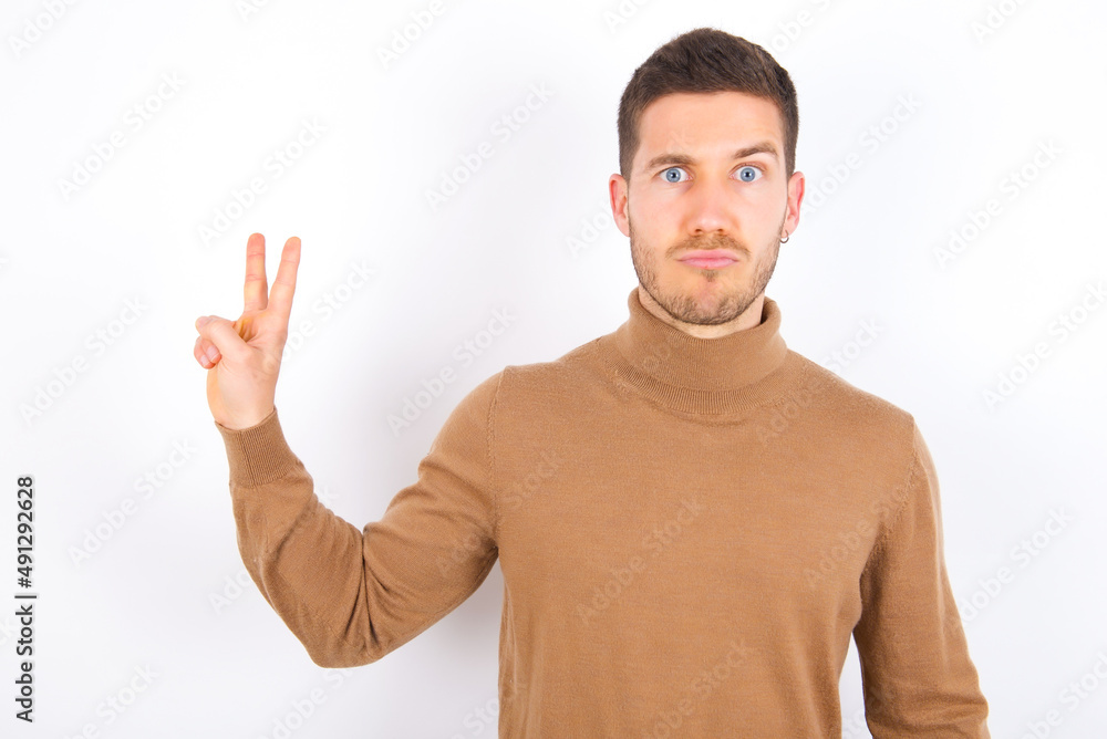 young caucasian man wearing knitted turtleneck over white background makes peace gesture keeps lips folded shows v sign. Body language concept