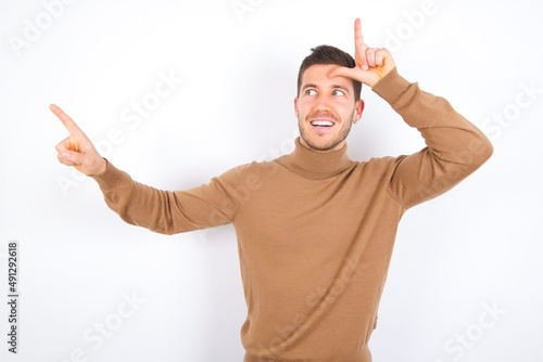 young caucasian man wearing knitted turtleneck over white background showing loser sign and pointing at empty space