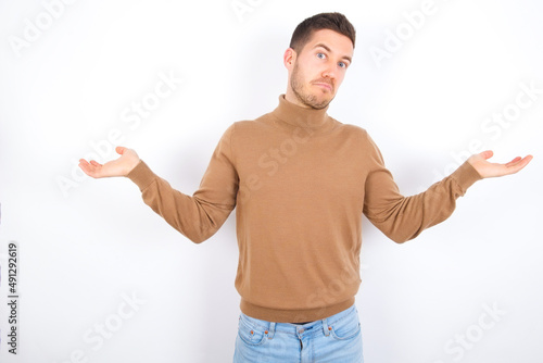 Careless attractive young caucasian man wearing knitted turtleneck over white background shrugging shoulders, oops.