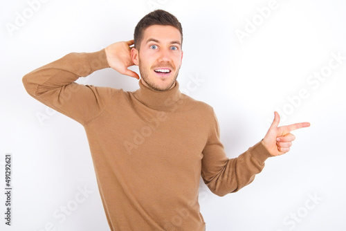 Surprised young caucasian man wearing knitted turtleneck over white background pointing at empty space holding hand on head