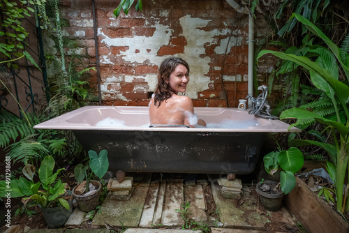 Valokuva A young woman looking over her shoulder smiles happily while sitting in an outdoor bubble bath in a lush tropical garden