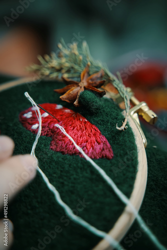 Process of embroidery of mushroom hat in wooden hoop on green material. Concept of needlework, hobby, leisure. Woman hands doing stitch.