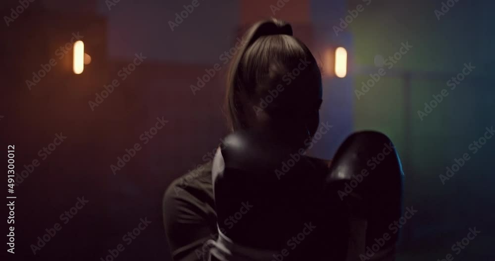 Portrait, a girl professionally practices punches in a dark hall of a fight club, boxing gloves on her hands. Silhouette, a girl boxer strikes in fighting gloves, a serious look of the face. Dark room Portrait, a girl professionally practices punches in a dark hall of a fight club, boxing gloves on her hands. Silhouette, a girl boxer strikes in fighting gloves, a serious look of the face. Dark room