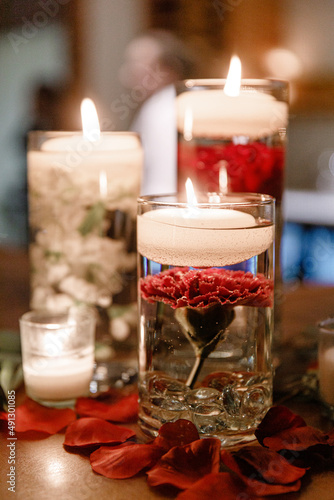 Lit candles in transparent glass jars on a wooden table A glass vases of water with burning candles and red flower inside Wedding table set decor Vases with red flowers Cropped photo Close up