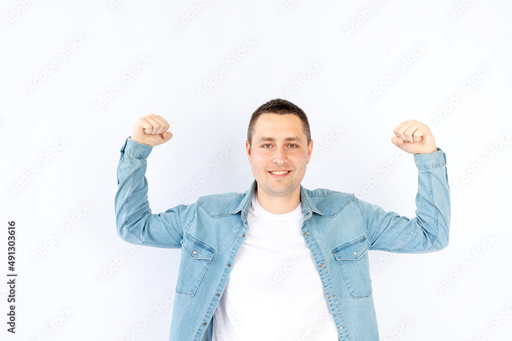 a young man or guy in a blue denim shirt on a white isolated empty background in a photo studio shows his strength by raising his hands up, empty space for text a young man or guy in a blue denim shirt on a white isolated empty background in a photo studio shows his strength by raising his hands up, empty space for text