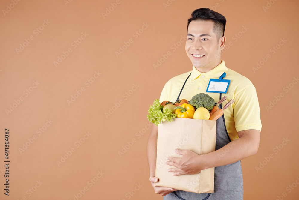 Happy Asian dark store worker carrying bag of fresh products collected for delivery to customer Happy Asian dark store worker carrying bag of fresh products collected for delivery to customer