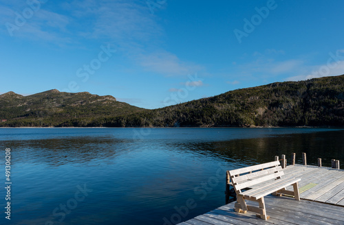 The view of two rolling hills covered in green trees from a wooden wharf with a bench on the deck of a wooden pier. The mountains are reflected in the still water. The sky is a vibrant blue with cloud
