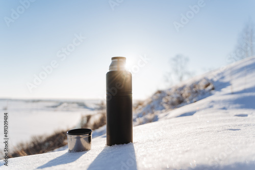 Sunlight glints with rays against the blue sky. A thermos of hot tea stands in the snow. Winter landscape, hiking in the forest, camping equipment.