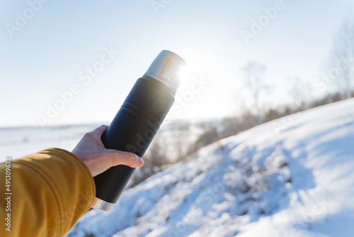 A hand holds a thermos of tea against the sky, sunlight glare into the camera. Black vacuum vessel bottle. The concept of winter trekking in the mountains.