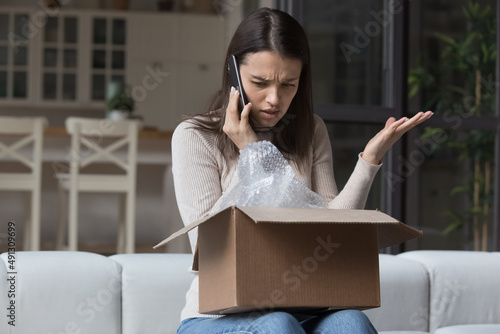 Dissatisfied angry woman, cheated client sit on sofa check received box, damaged or broken goods in parcel, talks to customers support, express complaints, looks annoyed. Bad delivery services concept