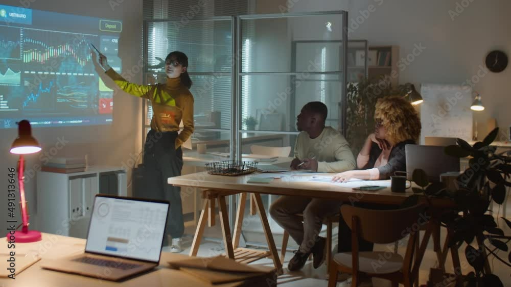 Young Asian businesswoman explaining financial data and graphs on projection screen to African American colleagues while having team meeting late at night in office Young Asian businesswoman explaining financial data and graphs on projection screen to African American colleagues while having team meeting late at night in office