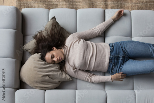 Overhead view tired young woman sleeping alone on couch at home. Peaceful female having deep daytime nap lying on cozy comfortable sofa in living room. Stress and exhaustion relieve, fatigue concept