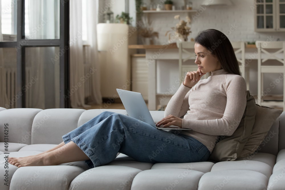 Pensive young woman sit on sofa staring at wireless computer screen feel concerned, looking thoughtful, working from home, search solution or ideas, choose goods online. Telecommute, challenge concept Pensive young woman sit on sofa staring at wireless computer screen feel concerned, looking thoughtful, working from home, search solution or ideas, choose goods online. Telecommute, challenge concept