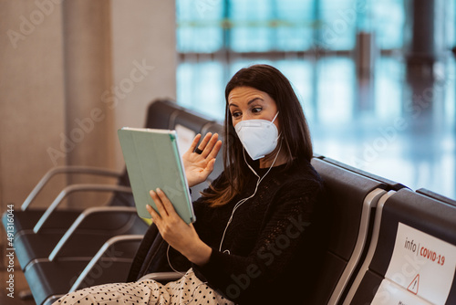 Tourist woman at the airport waiting for her flight. Wearing a mask for covid 19 protection doing a videocall with her tablet. Lifestyle. Travel
