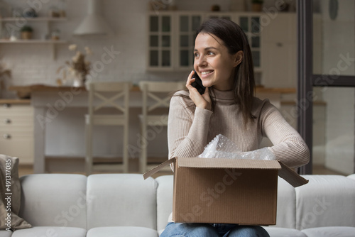 Smiling young woman sits on sofa at home with cardboard parcel, using smartphone talking to friend, express gratitude for received gift in box. E-commerce client, satisfied shopper, delivery concept photo
