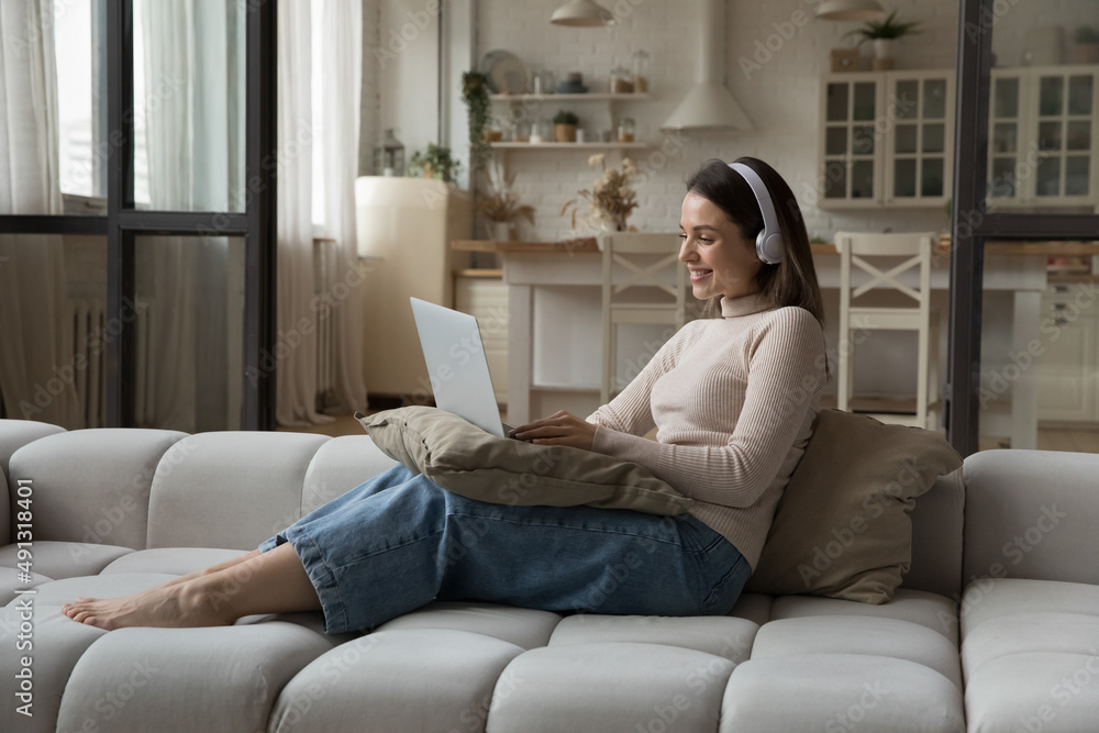 Smiling attractive young woman in headphones use laptop seated on cozy sofa in living room. Makes video call, listens audio course, watch amusing video online, spend leisure on internet. Tech concept Smiling attractive young woman in headphones use laptop seated on cozy sofa in living room. Makes video call, listens audio course, watch amusing video online, spend leisure on internet. Tech concept