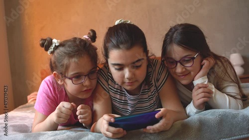 Three little female friends were lying on the bed ,talking and laughing.They use a smart phone to play games.	
