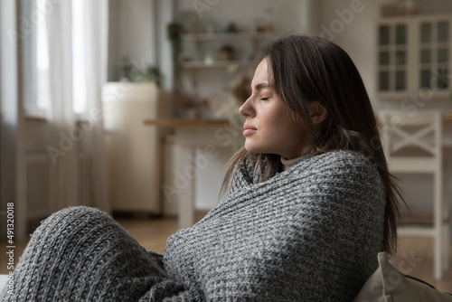 Картината върху платно Young woman covered in warm knitted plaid sits on sofa at home suffers from low temperature in living room, shivers, having flu symptoms feeling unwell
