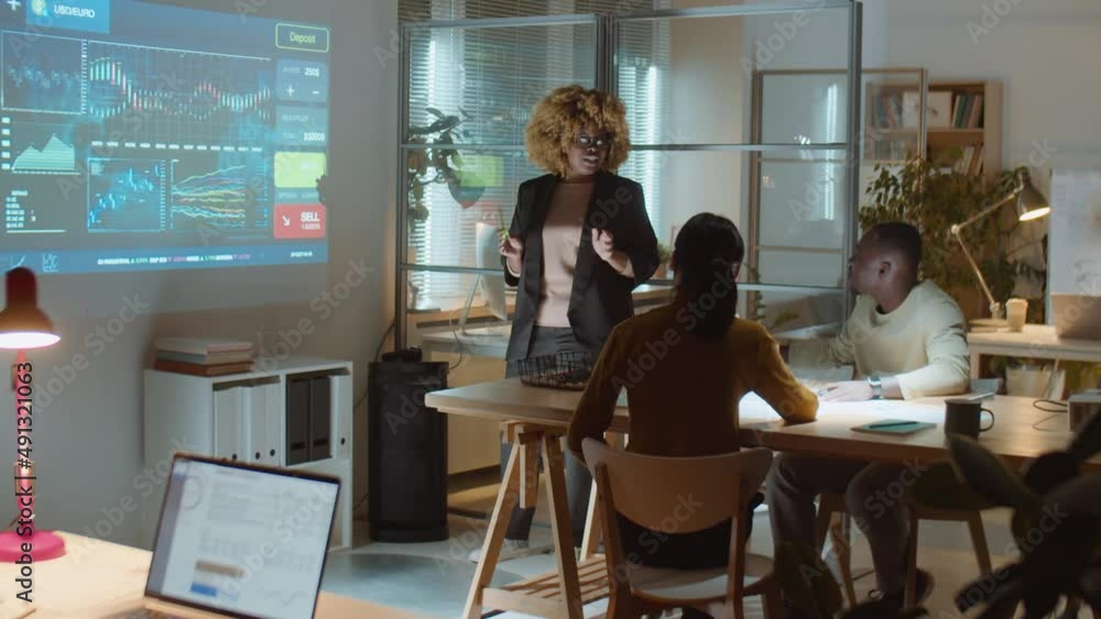 African American businesswoman explaining financial data on projection screen while giving presentation to team during night office meeting African American businesswoman explaining financial data on projection screen while giving presentation to team during night office meeting