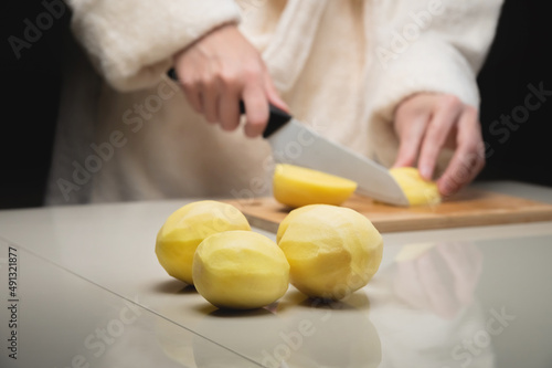 Woman in bathrobe cutting raw peeled potatoes on cutting wooden board at home kitchen