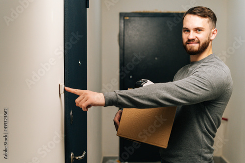 Delivery man holding cardboard box and contactless payment POS wireless terminal for card paying and ringing doorbell of customer apartment, looking at camera. Concept of online shopping.