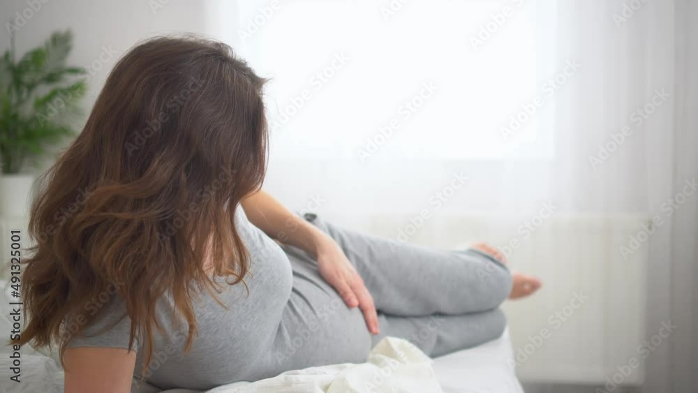 A cute pregnant woman sits in bed and holds her hand on her belly. The concept of a happy pregnancy in anticipation of a baby. 9 months of waiting for a baby. A cute pregnant woman sits in bed and holds her hand on her belly. The concept of a happy pregnancy in anticipation of a baby. 9 months of waiting for a baby.