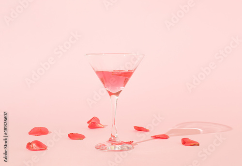 Cocktail glass and red flower petals with shadow on pink background. MInimal spring or season party concept.