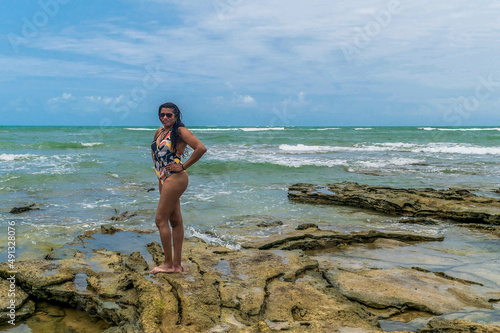 Mulher em linda praia, com céu azul e nuvens ao fundo, grandes pedras ao redor e ondas Trancoso, Bahia. © Diovane