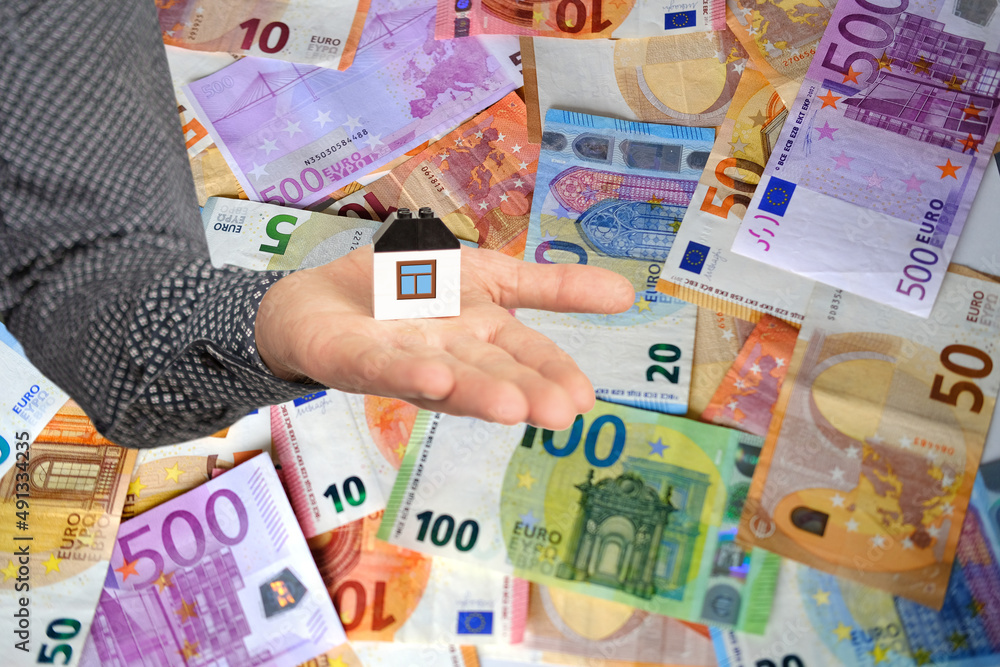 close-up of male hand holds model of house on lot of money background, paper euro banknotes, concept of cash, payments, savings, banking, save up to buy house, purchase real estate in mortgage, loan