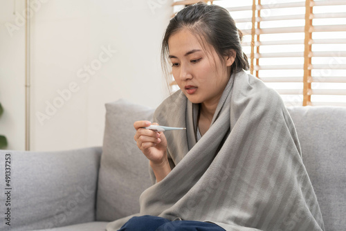 Sick, influenza asian young woman, girl headache have a fever, flu and check thermometer measure body temperature, feel illness sitting on sofa bed at home. Health care person on virus, covid-19.