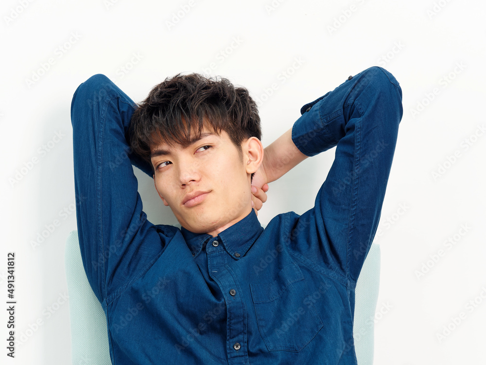 Portrait of handsome Chinese young man with curly black hair in blue shirt sitting in armchair and posing against white wall background. Hands behind head and looking away, front view studio shot. Portrait of handsome Chinese young man with curly black hair in blue shirt sitting in armchair and posing against white wall background. Hands behind head and looking away, front view studio shot.