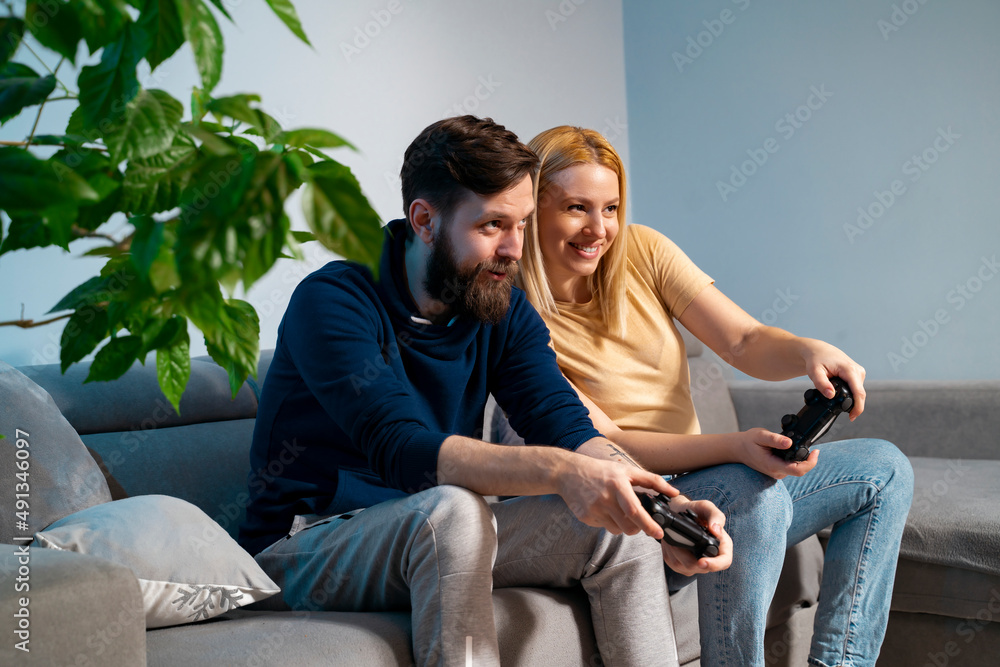 Man and woman enjoying playing video games at home on sofa. Blonde Caucasian woman and breaded man with game controllers in hands Man and woman enjoying playing video games at home on sofa. Blonde Caucasian woman and breaded man with game controllers in hands