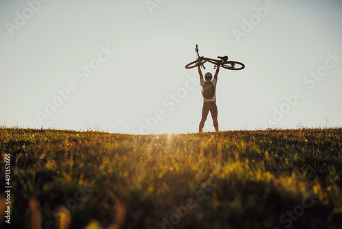 Cyclist is holding his bicycle over himself on the background of red sunset. Biker with bicycle on the field during sunrise