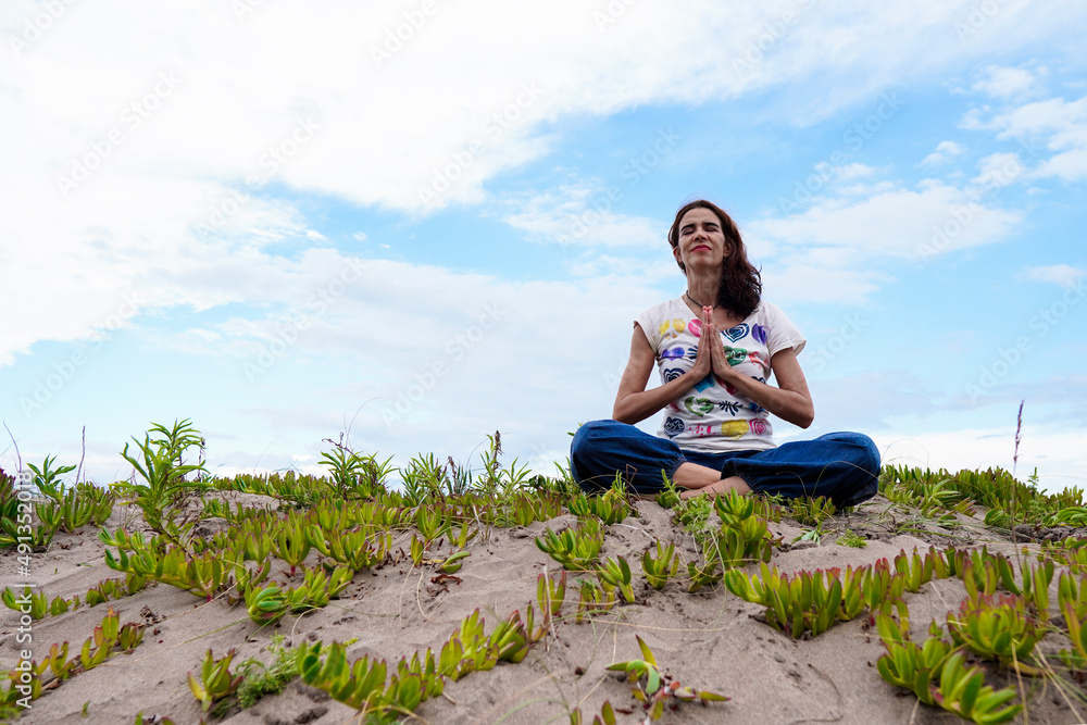 Adult latin woman smiling and doing yoga in the dunes receiving the spring season in a praying pose and close eyes, feeling the fresh air. Freedom and joyful lifestyle concept.