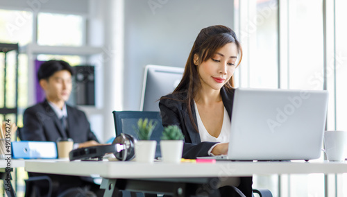 Asian young female professional businesswoman secretary employee in formal suit sitting at working desk typing report via laptop notebook computer while businessman manager work behind in office