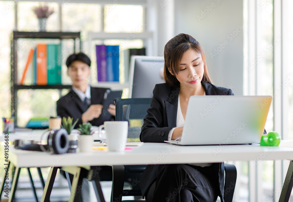 Asian young female professional businesswoman secretary employee in formal suit sitting at working desk typing report via laptop notebook computer while businessman manager work behind in office Asian young female professional businesswoman secretary employee in formal suit sitting at working desk typing report via laptop notebook computer while businessman manager work behind in office