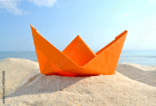 Bright orange paper boat in yellow sand on background of sea waves and blue sky on sunny summer day. Colored ship close-up. Concept adventures travelling vacation holiday rest tourism voyager dream