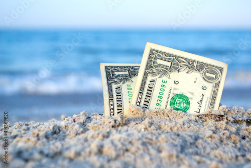 Two one dollar bills half buried in sand on sandy seashore close-up. Two paper dollar bills half in sand on sea beach on sunny day. Concept money, finance, investment, wealth, poverty, financing, cash