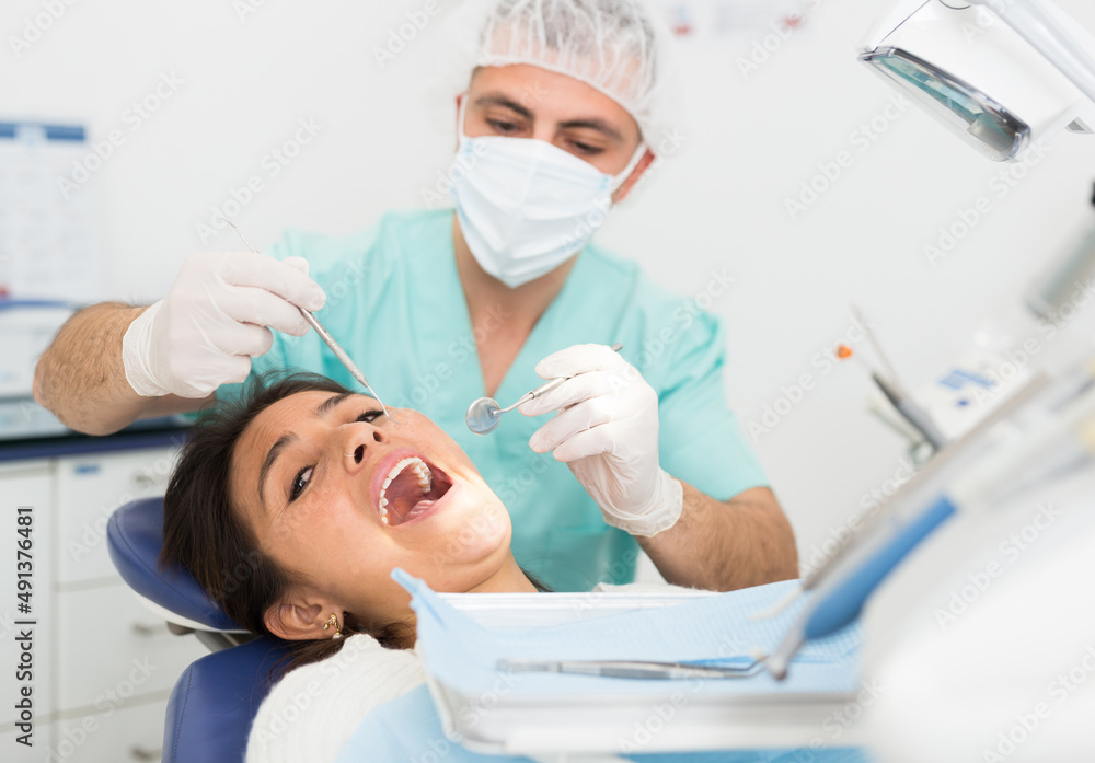 Fototapeta premium Dentist man examining a latin female patient teeth with dental tools - mirror and probe