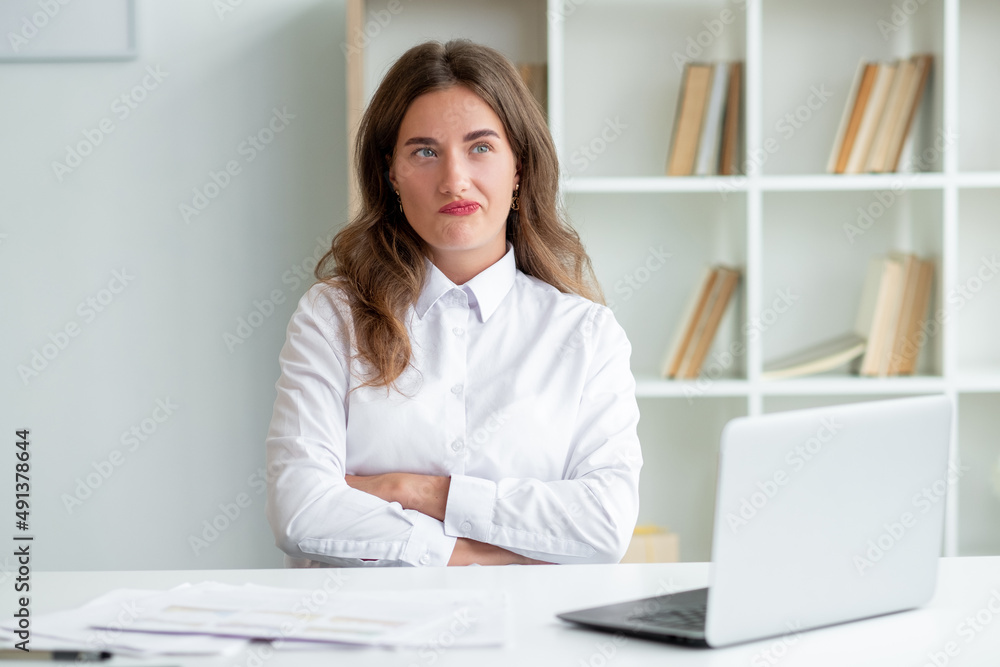 Consider problem. Skeptic woman. Puzzled emotion. Displeased lady crossing hands on chest sitting desk with documents and opened laptop in light room interior. Consider problem. Skeptic woman. Puzzled emotion. Displeased lady crossing hands on chest sitting desk with documents and opened laptop in light room interior.