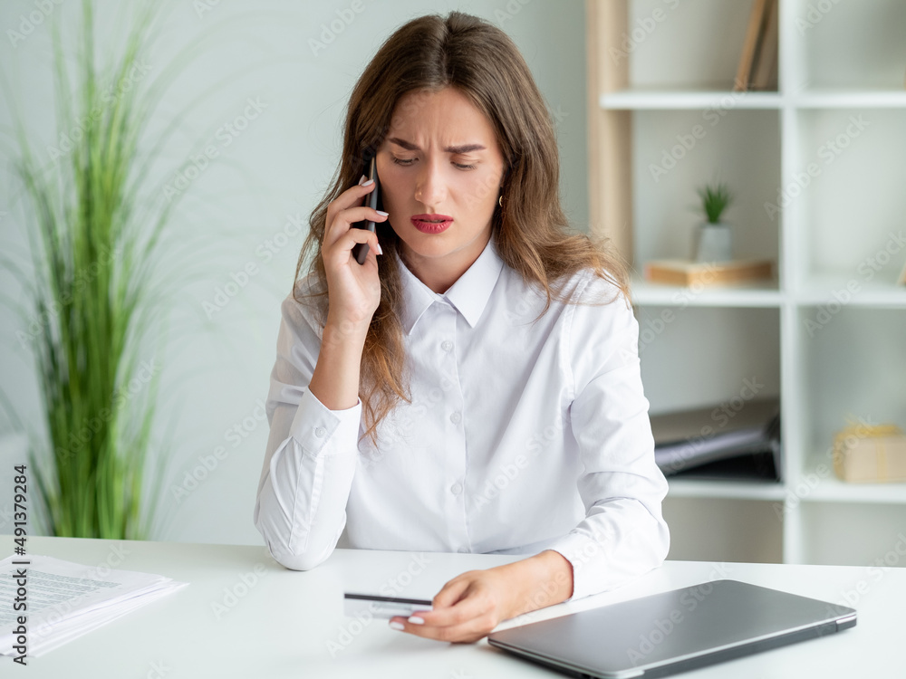 Bank payment. Worried woman. Mobile consultation. Considered office lady holding credit card talking smartphone sitting work desk in light room interior. Bank payment. Worried woman. Mobile consultation. Considered office lady holding credit card talking smartphone sitting work desk in light room interior.