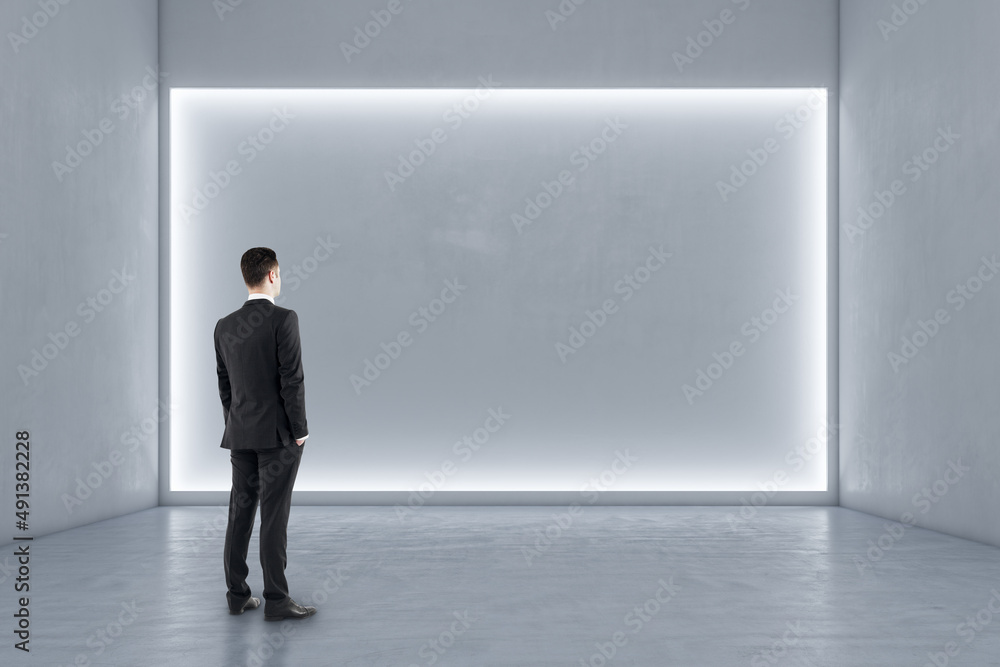 Businessman standing in modern exhibition hall interior with illuminated white mock up wall and concrete floor. Gallery concept. Businessman standing in modern exhibition hall interior with illuminated white mock up wall and concrete floor. Gallery concept.