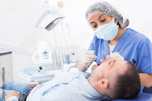 Doctor dentist woman in face mask examining a male patient teeth with dental tools